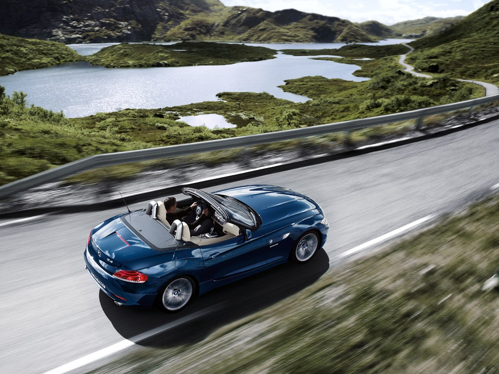 Mavi BMW Z4 E89 açık tavanla göl kıyısındaki yolda arkadan üst açı