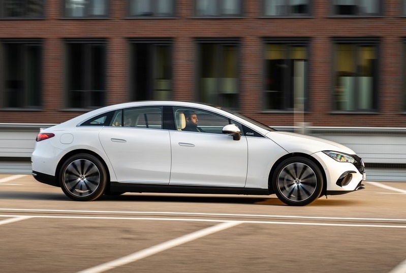 Beyaz Mercedes-Benz EQE sedanın şehir içinde hareket halindeki yan profili