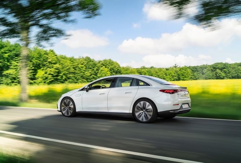 Beyaz Mercedes-Benz EQE sedanın ağaçlıklı yolda hareket halindeki arka yan görünümü